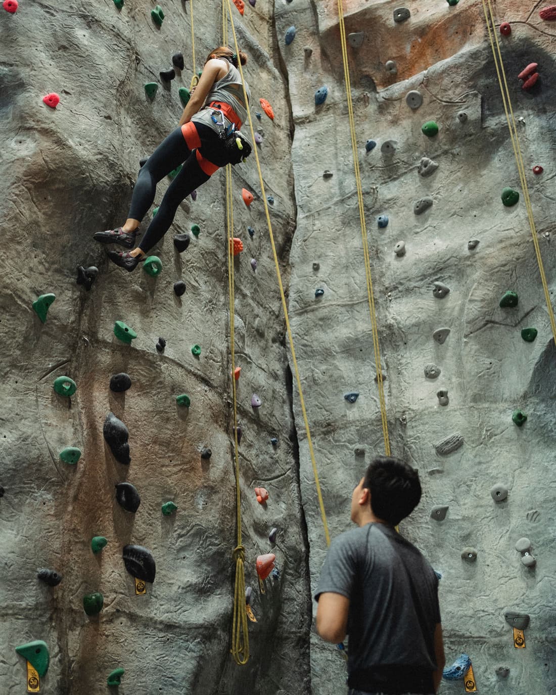 A climber part-way up a top-rope route on an indoor climbing wall, watched from the ground by a belayer holding the rope.