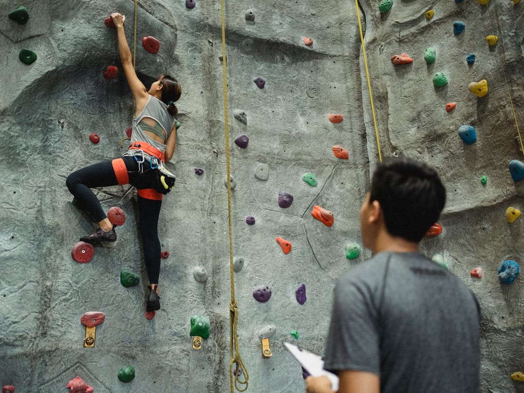 A climber part-way up an indoor top-rope wall marked with route grades, watched from the ground by an instructor holding a clipboard.