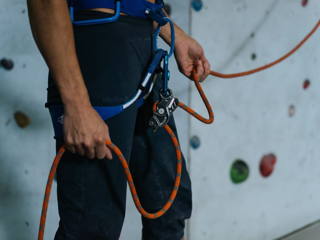 A belayer at an indoor wall with the rope running through a belay device clipped to the harness, one hand on the rope above the device and the braking hand below it.