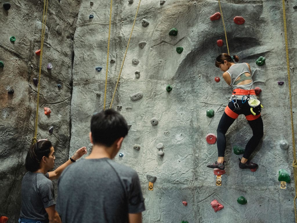 A wide view of an indoor climbing wall with two people watching from the ground as a climber works up a graded route.